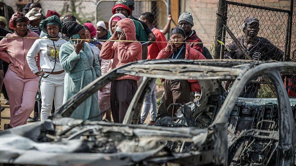 Crowd around a burned out car outside a bar in Johannesburg after attack.