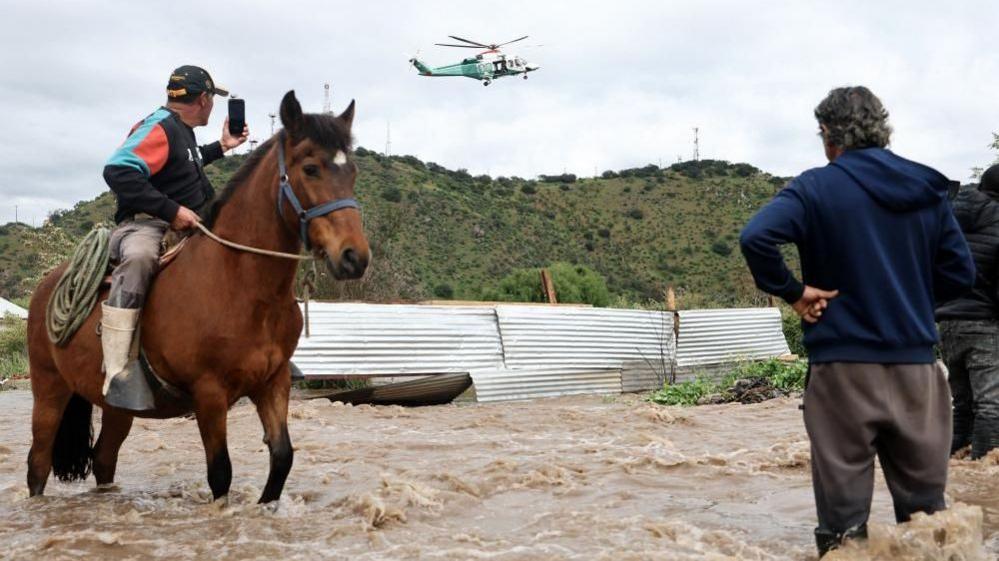Man on horse filming helicopter overhead in flooded water, Talagante, Chile