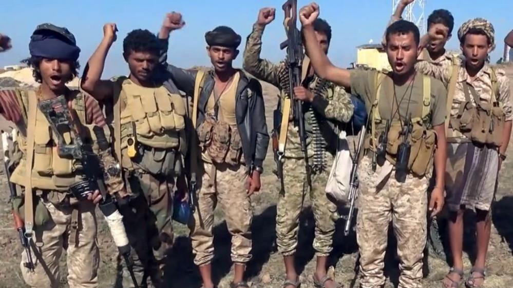 Houthi fighters wearing camouflage uniforms and guns strapped to their shoulders raise fists in the air