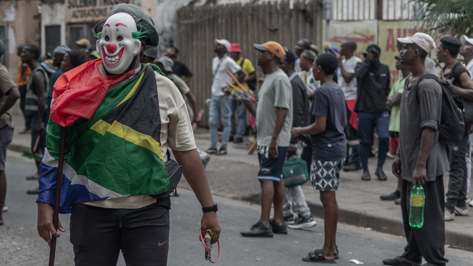 Mask‑d man draped in South African flag stands in the street as onlookers gather behind him