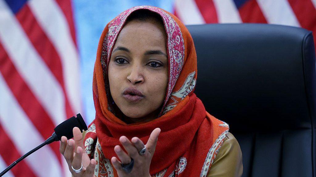 Ilhan Omar in a black chair with an orange headcover