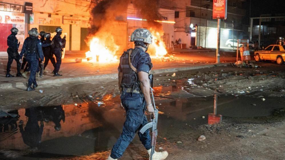 Police officers patrol a street as burning tyres and debris send flames and thick smoke into the air during protests over prolonged power cuts